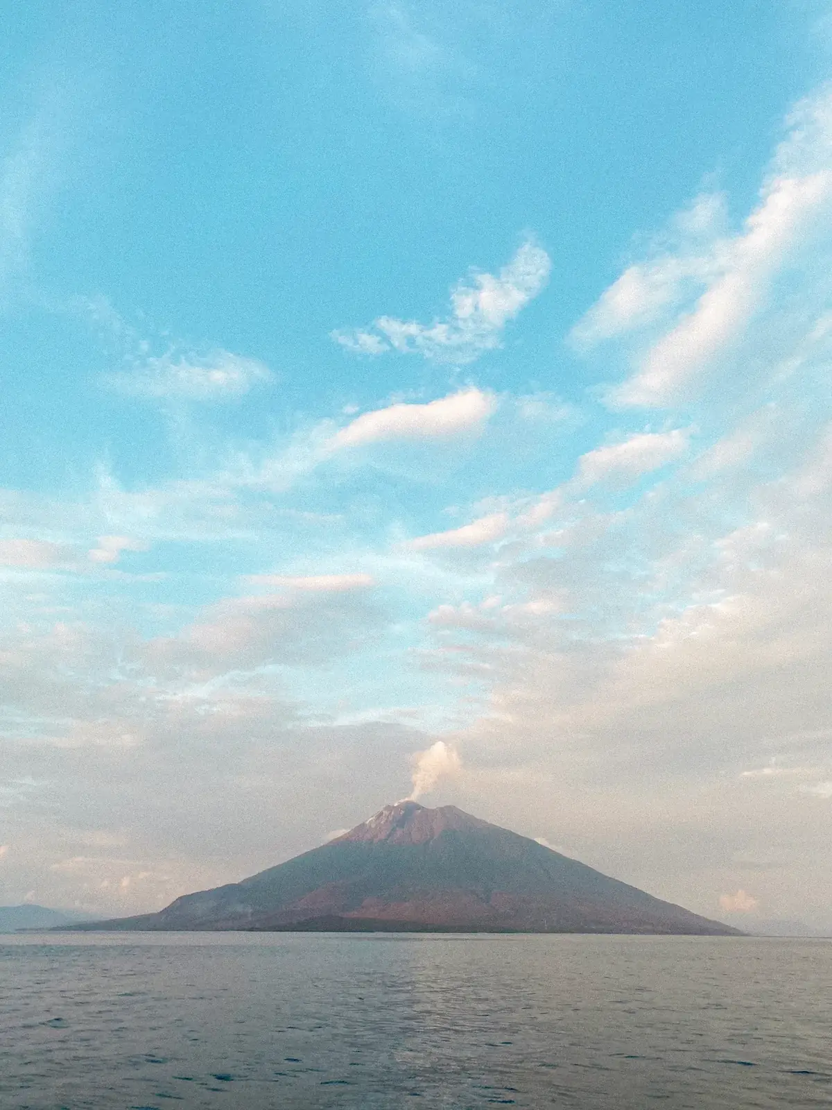 A cone volcano rising from the sea under a pastel sky.