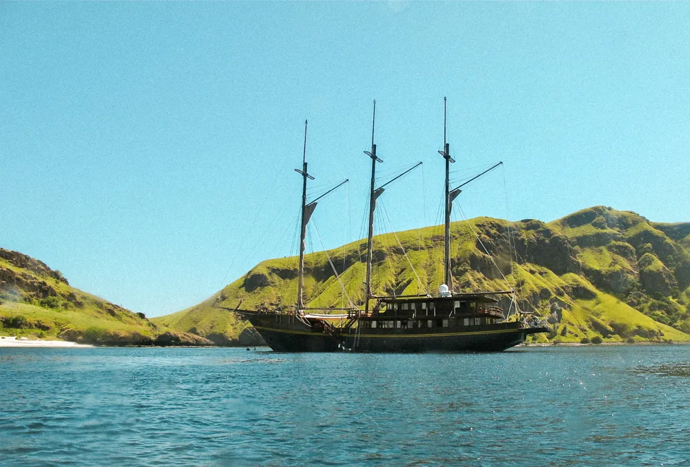 A three-masted ship anchored below green hills.