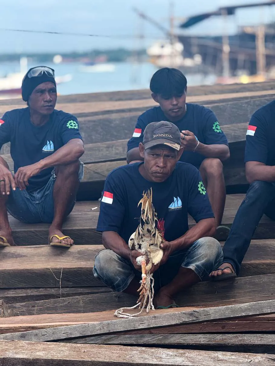 Shipyard workers seated on timbers, one holding a rooster