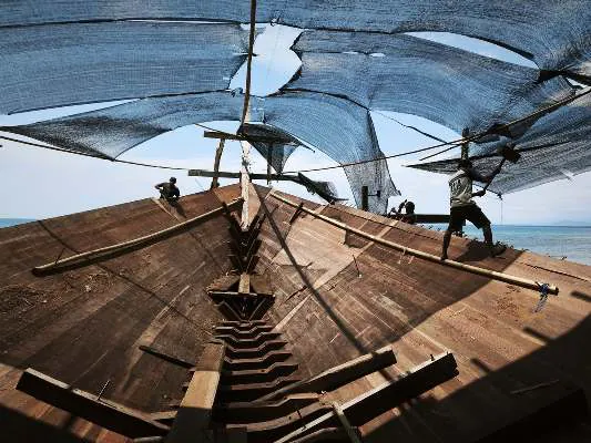 A shipwright working on the bow of WAOW 2 under shade netting, the sea behind