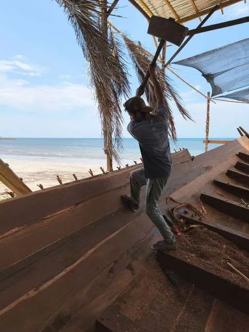 A shipwright driving a plank into place with a mallet, the beach behind