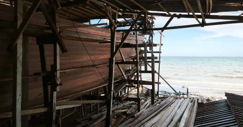 The planked hull on its scaffolding by the sea