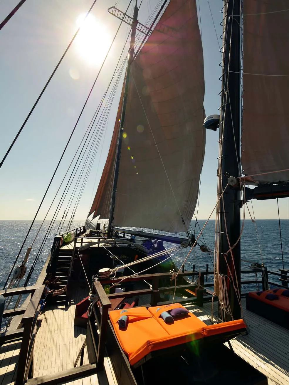 The deck of a phinisi under sail with orange cushions, sun shining through the sails