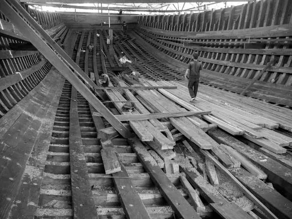 Black-and-white view along the inside of the hull, a worker walking on the planks