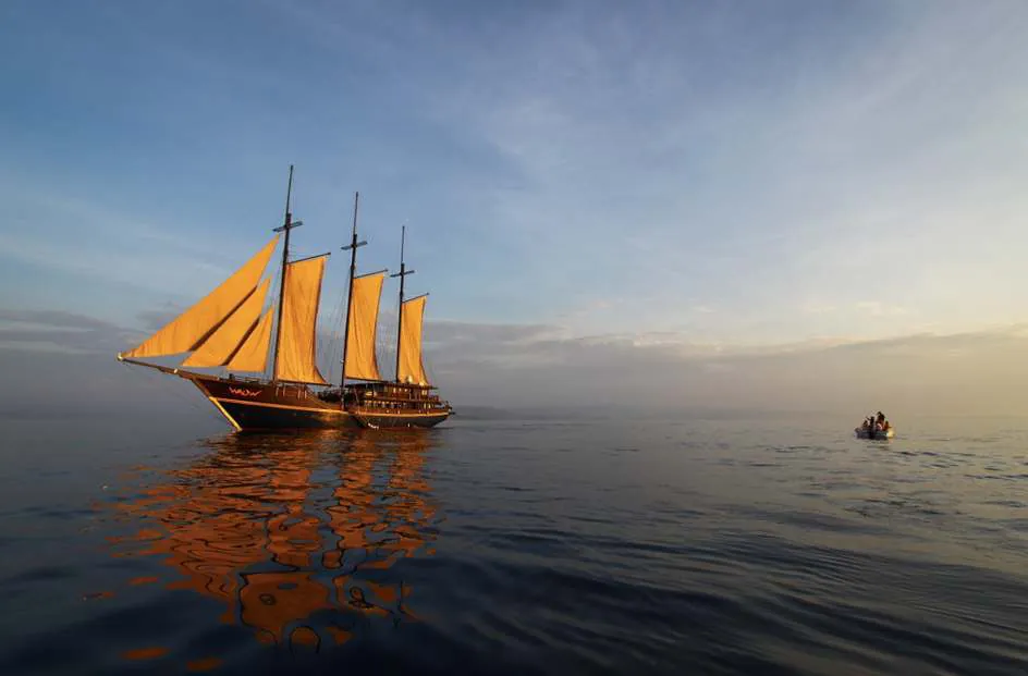 A three-masted phinisi with golden sails on a calm sea at sunset, a small tender nearby