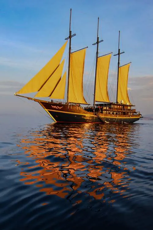 A phinisi under full golden sail at sunset, reflected in a glassy sea