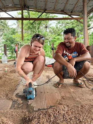 Camille using a power sander on a plank next to a smiling craftsman
