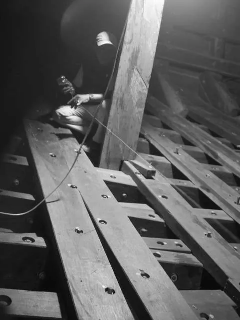 Black-and-white photo of a craftsman working by lamplight inside the hull