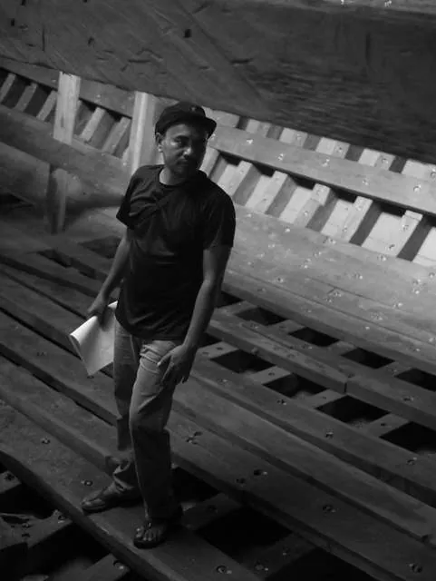 Black-and-white photo of a man holding plans, standing among the frames inside the hull