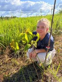A woman crouching next to a young planted tree in tall grass