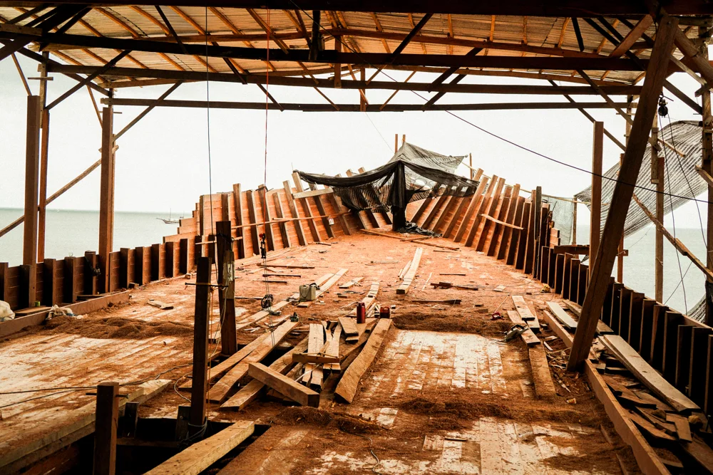 A wooden phinisi hull under construction in a shed.
