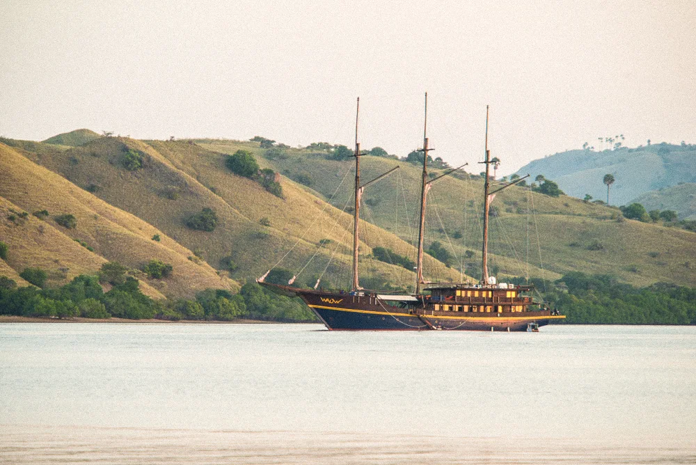 A three-masted phinisi anchored below dry hills.